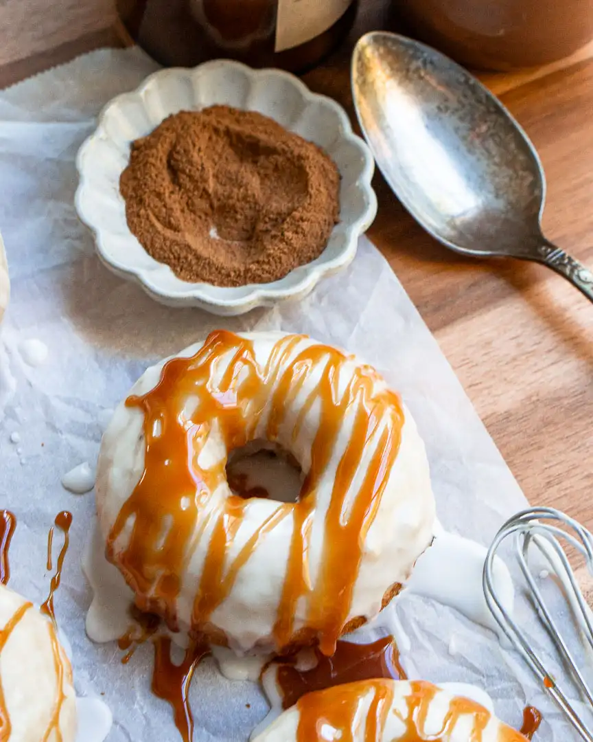 A vanilla frosted donut drizzled with a caramel glaze. The donut is sitting next to a small bowl of ground cinnamon and a spoon.