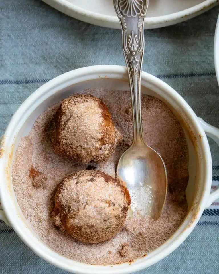 Two pumpkin bites covered in cinnamon sugar. They are in the bowl of cinnamon sugar and there is a spoon to help coat the donuts.