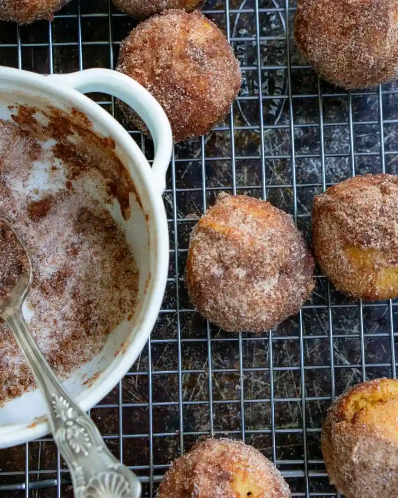 Pumpkin bites coated with cinnamon sugar and on a cooling rack. There is a bowl of cinnamon sugar next to the donuts.