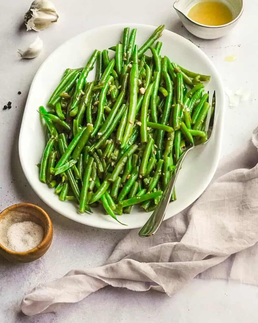 Platter of cooked green beans with olive oil pitcher, salt and napkin next to serving dish.