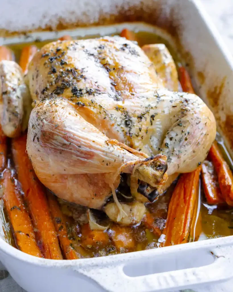 Roasted whole chicken a bed of carrots in a baking pan with pan juices.