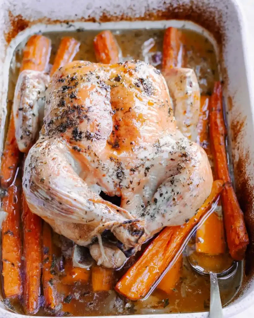 Roasted whole chicken on a bed of carrots in a roasting pan.