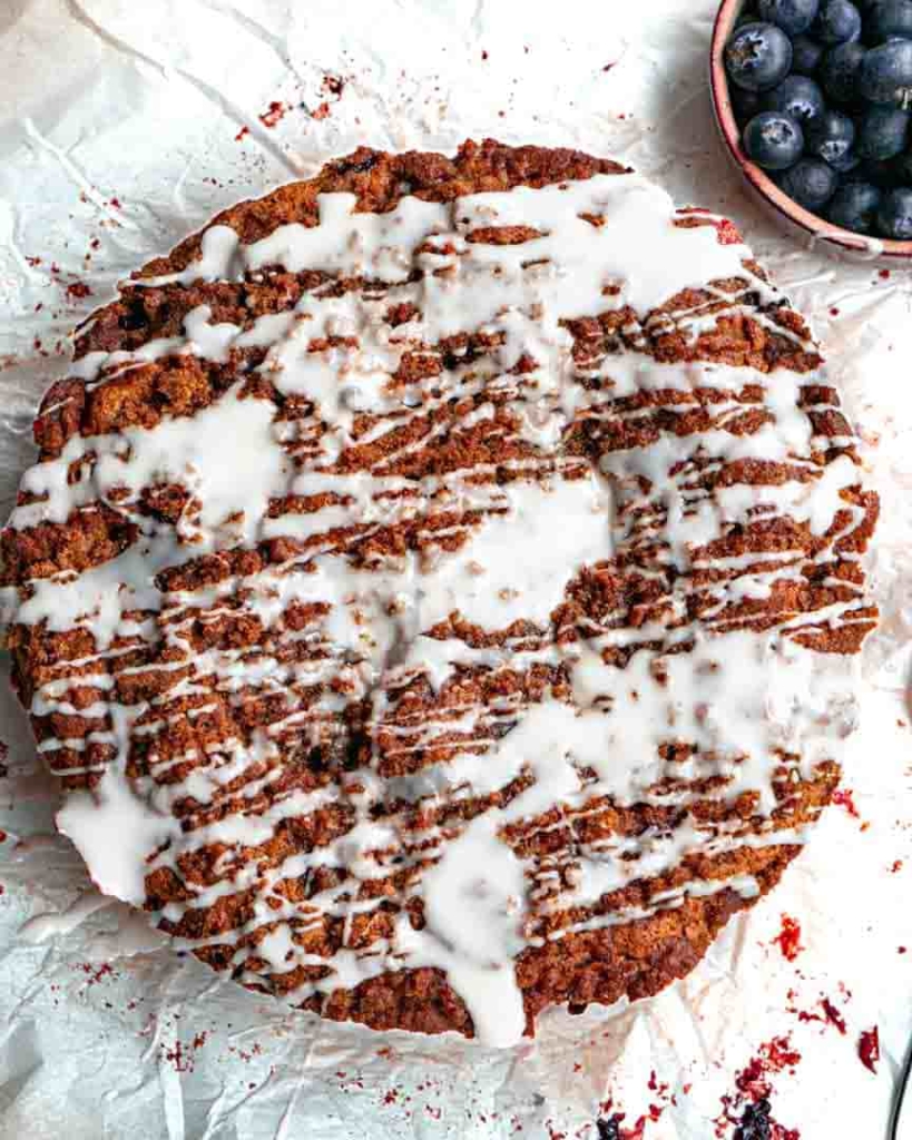 A round blueberry coffee cake with glaze drizzled on top. There is a small bowl of blueberries next to the cake.