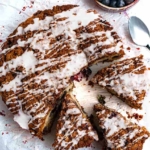 A round gf blueberry streusel coffee cake that has some cut pieces pulled away from the cake. There is a spoon next to the cake that has some glaze on it. There is a small bowl of blueberries next to the cake.