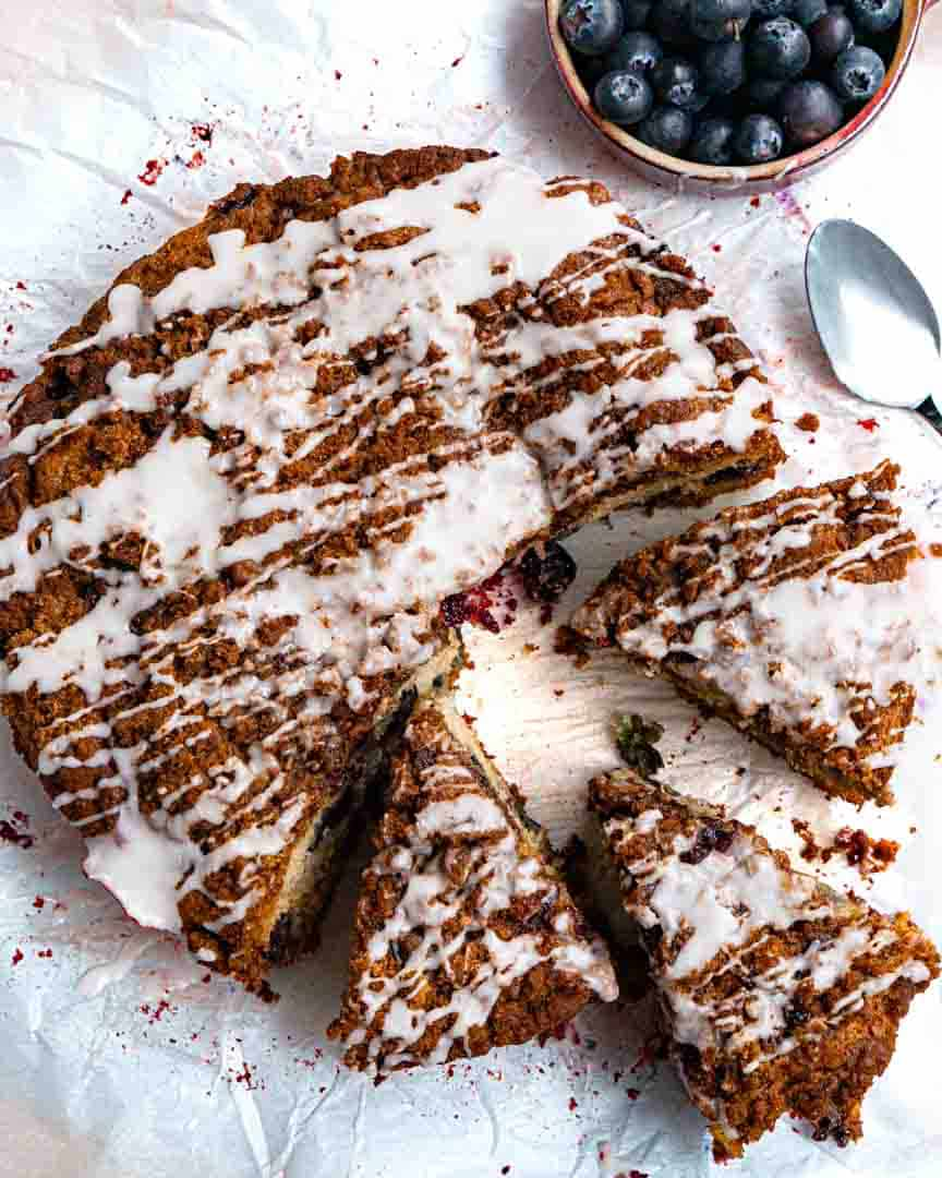 A round gf blueberry streusel coffee cake that has some cut pieces pulled away from the cake. There is a spoon next to the cake that has some glaze on it. There is a small bowl of blueberries next to the cake.