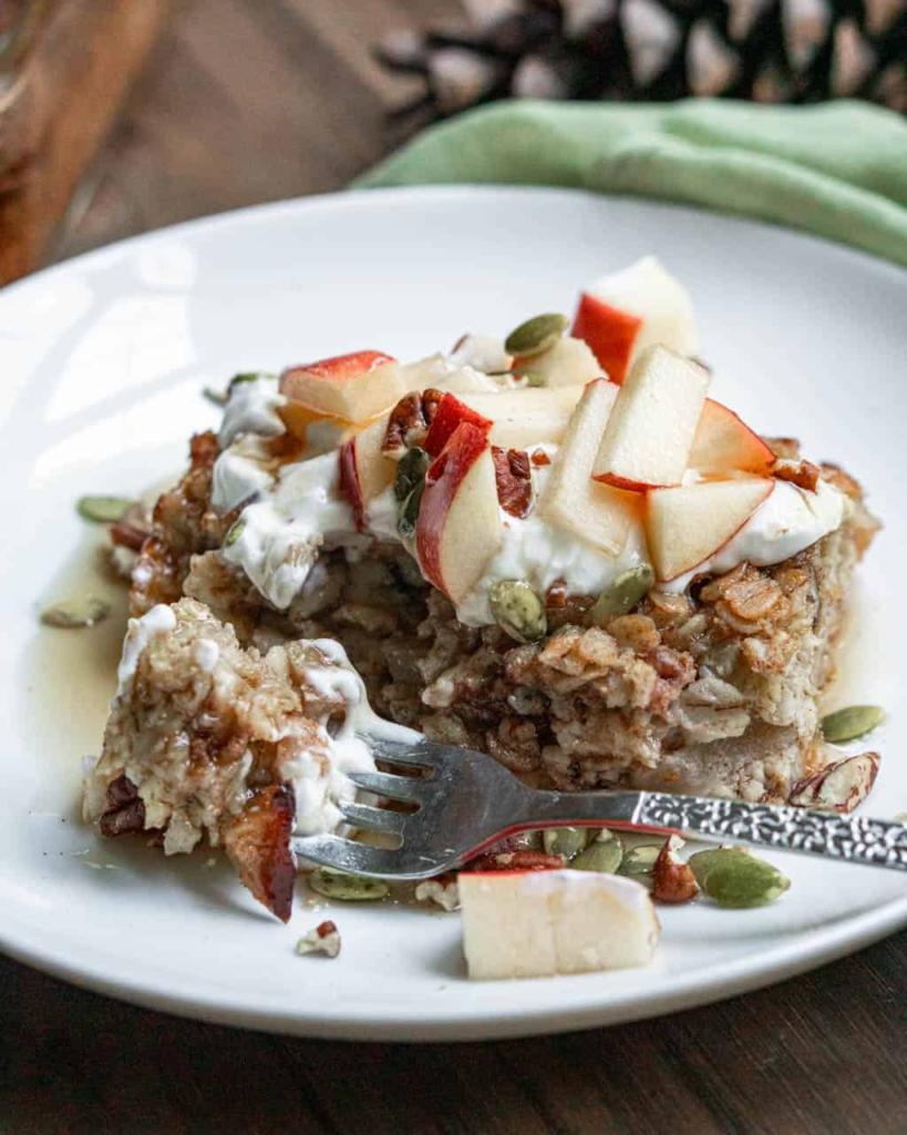 A piece of baked oatmeal topped with yogurt, sliced fresh apples, and pumpkin seeds on a plate with a fork