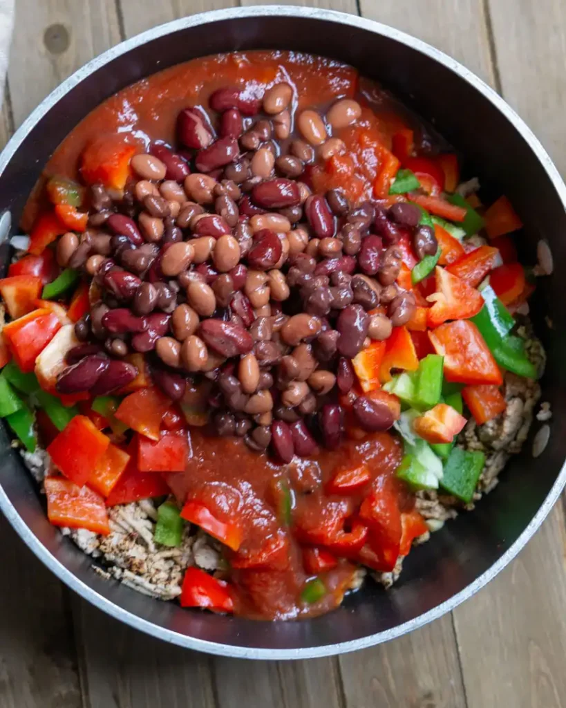 A pot of cooked ground turkey with chopped raw red and green peppers, chopped onions and a can of mixed beans on top of the ground turkey.