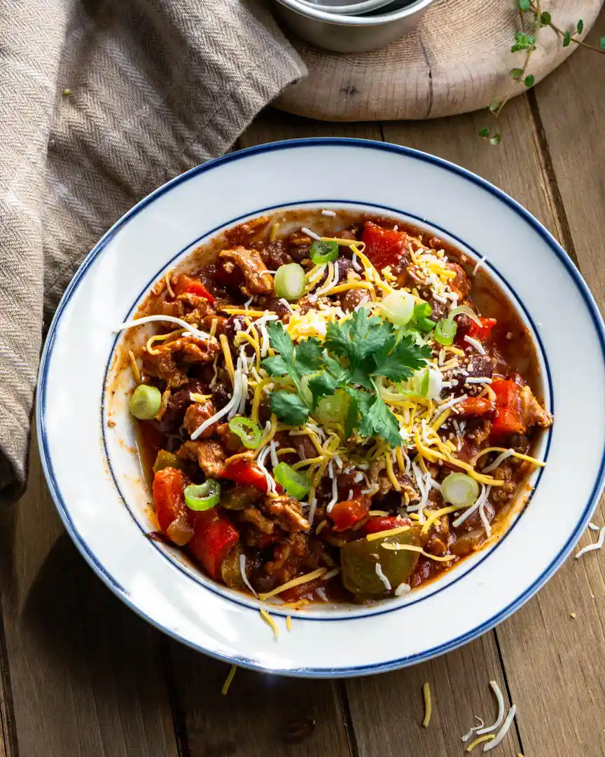 A bowl of chili with shredded cheese, sliced green onions and cilantro on top of it.