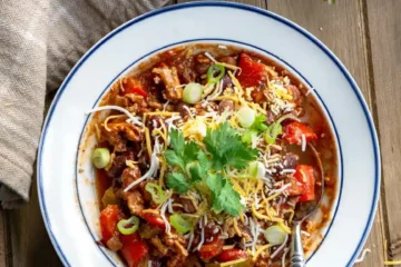 A bowl of chili with shredded cheddar cheese, sliced green onions, and cilantro on top.