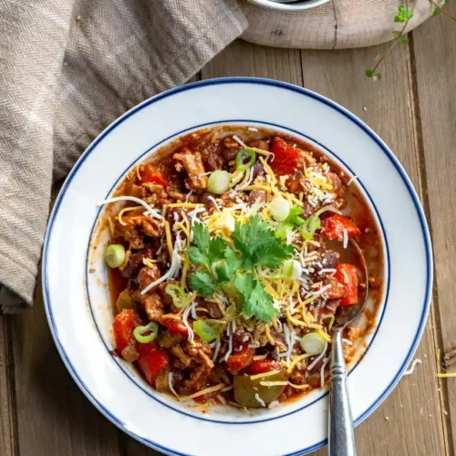 A bowl of chili with shredded cheddar cheese, sliced green onions, and cilantro on top.