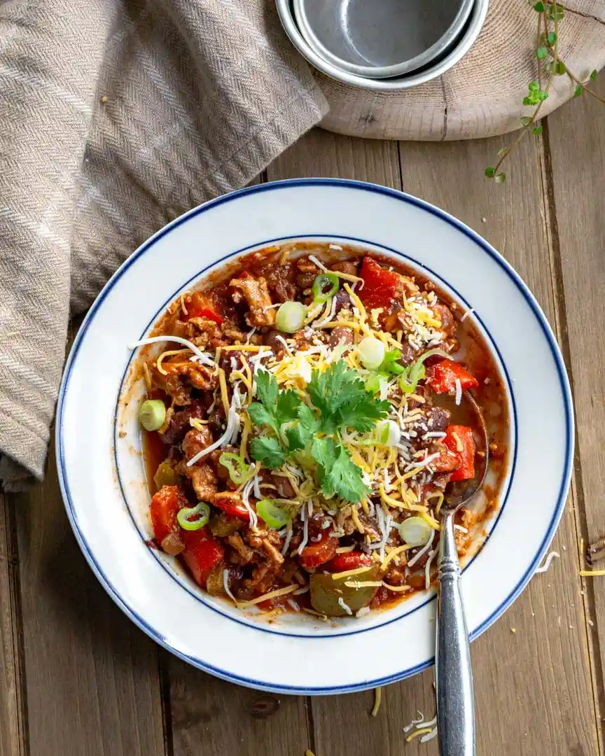 A bowl of chili with shredded cheddar cheese, sliced green onions, and cilantro on top.