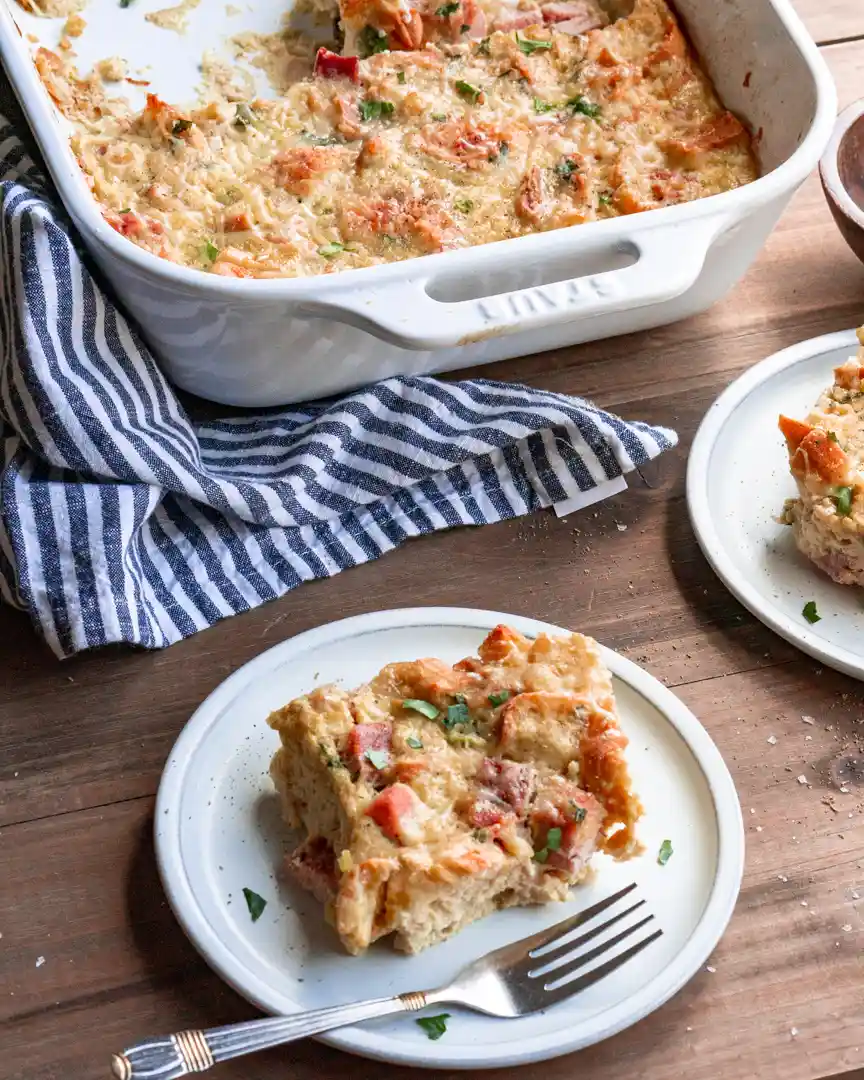 cut piece of strata on a plate with the baking pan filled with some strata next to it.