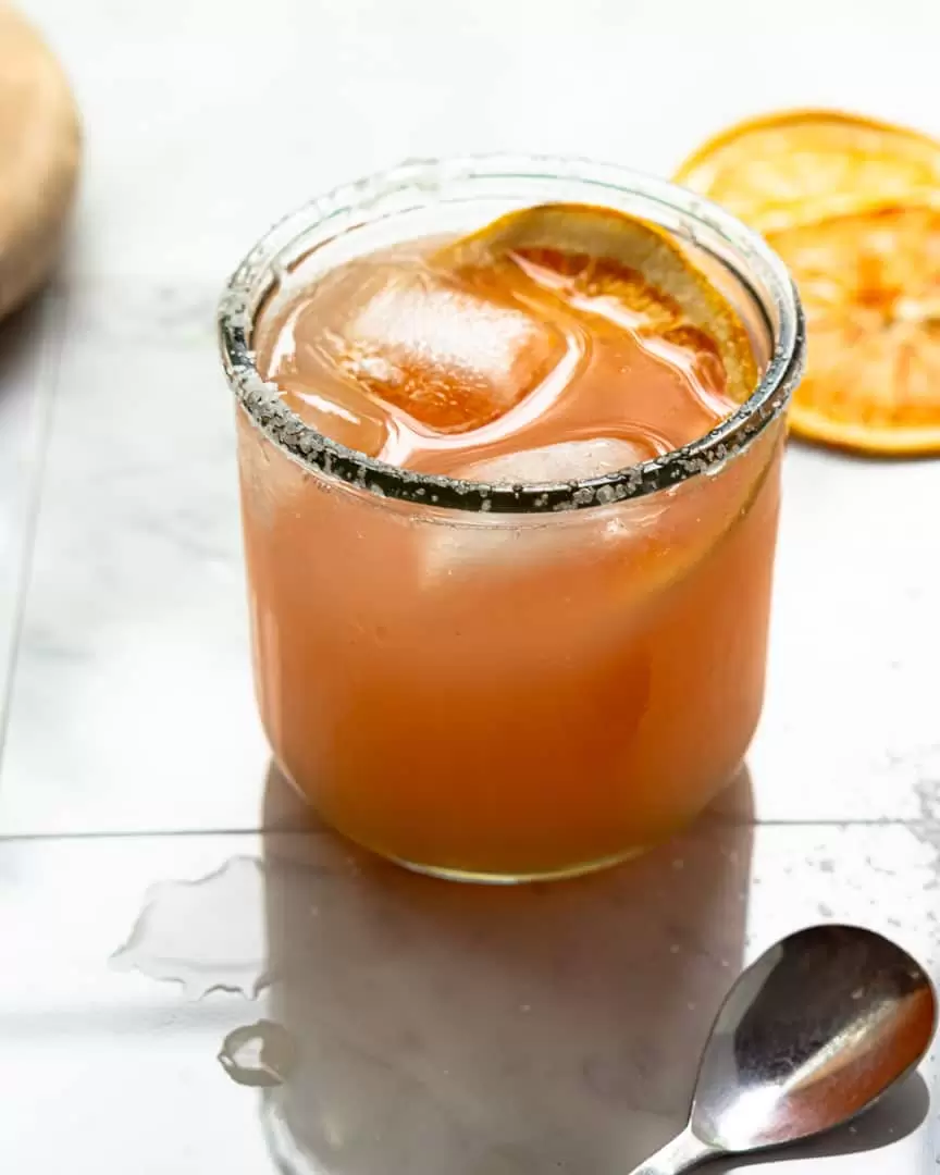 Paloma cocktail in a short glass with ice cubes and dried orange slice floating on top. With dried orange slices and a spoon next to the glass.