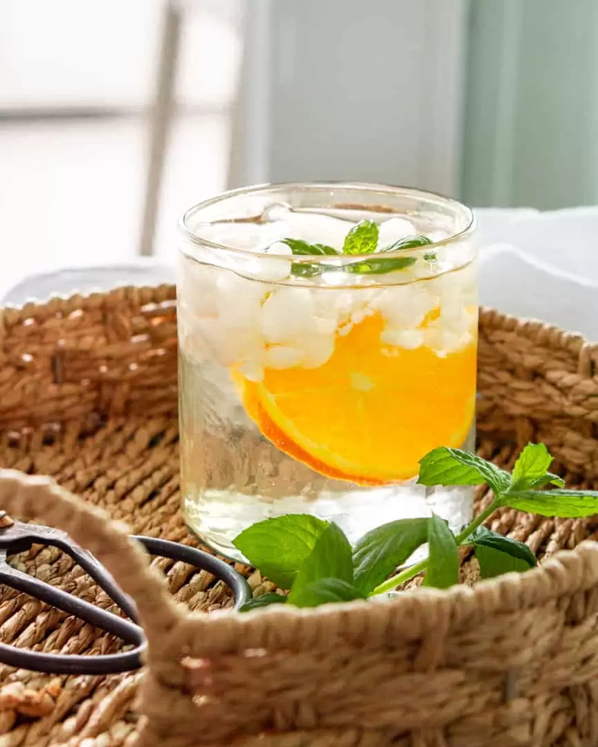 glass of ice water with a slice of orange and a sprig of mint on a wicker tray