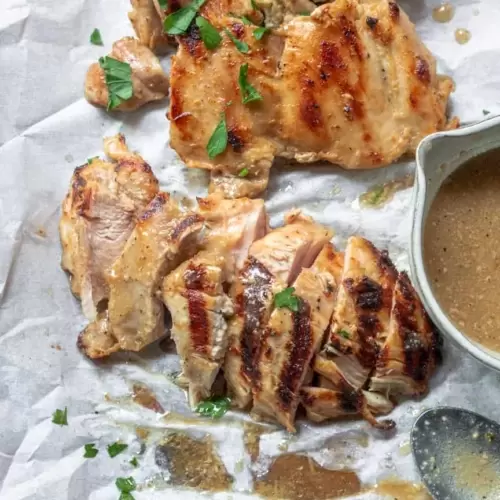 chicken thighs sliced on a baking sheet with a spoon next to it and a small bowl of dressing.