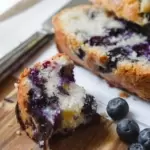 A small broken piece of blueberry loaf next to sliced cake and fresh blueberries.