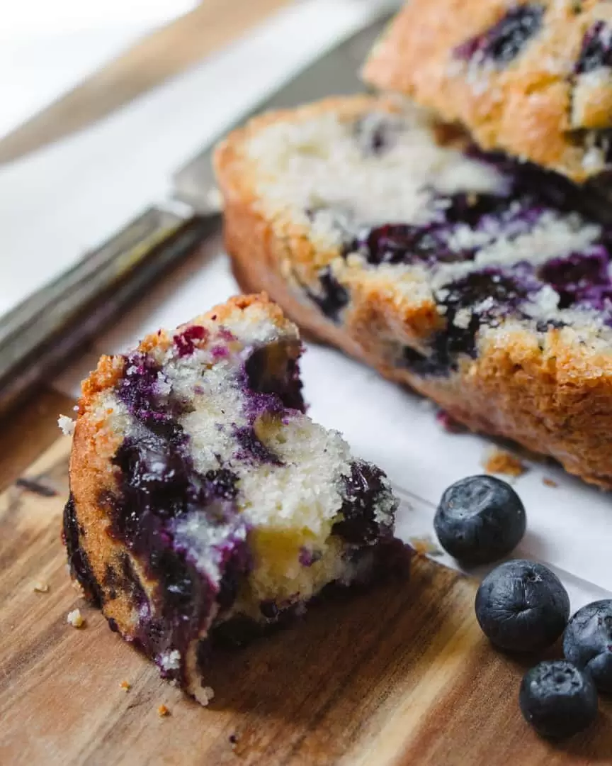 A small broken piece of blueberry loaf next to sliced cake and fresh blueberries.