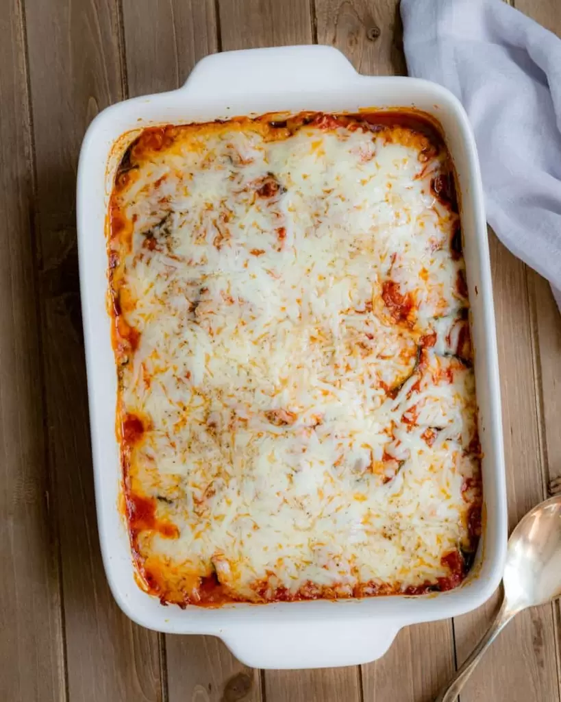 A casserole dish with layered zucchini, sauce and topped with mozzarella cheese. A serving spoon next to the casserole pan.