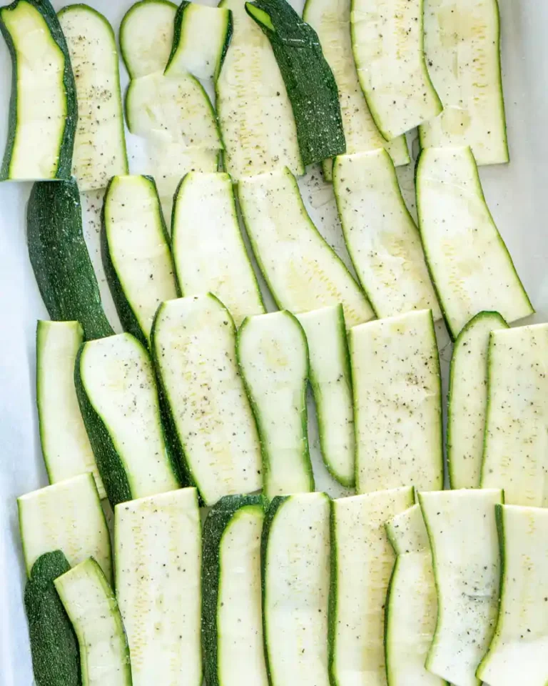 Raw zucchini sliced vertically and slightly layered onto a baking sheet with salt, pepper and olive oil drizzled on top.