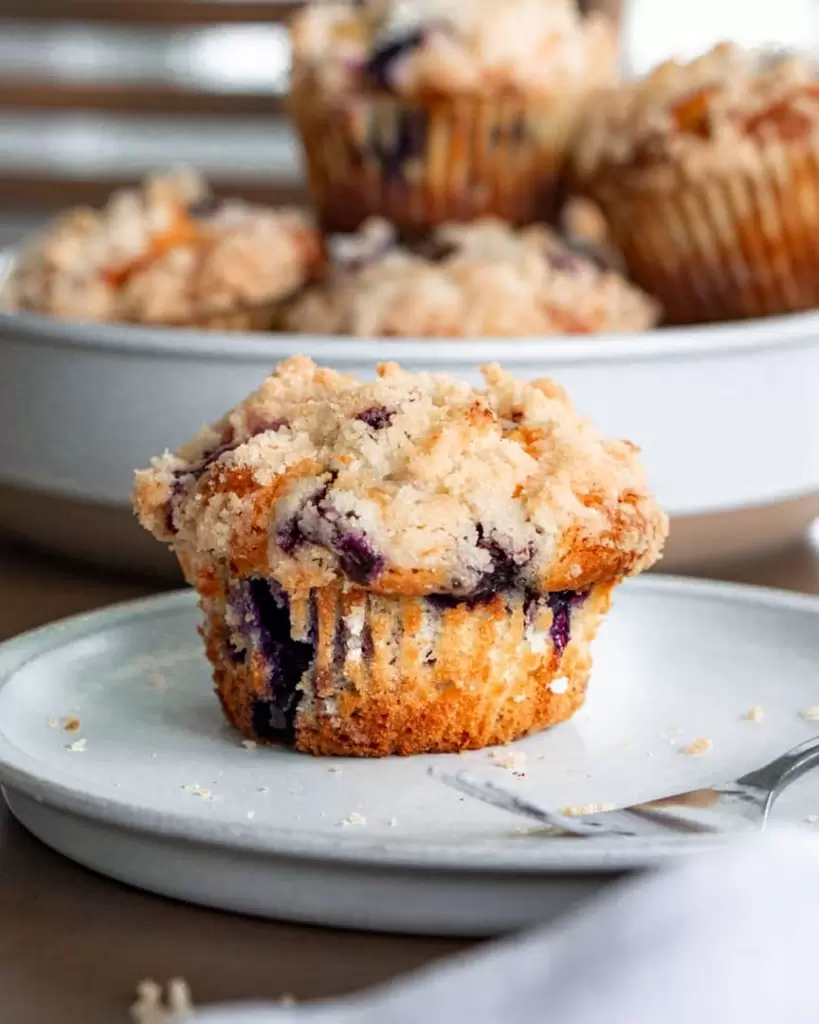 Blueberry muffin on a plate with a fork next to it. A plate of blueberry muffins stacked in the background.