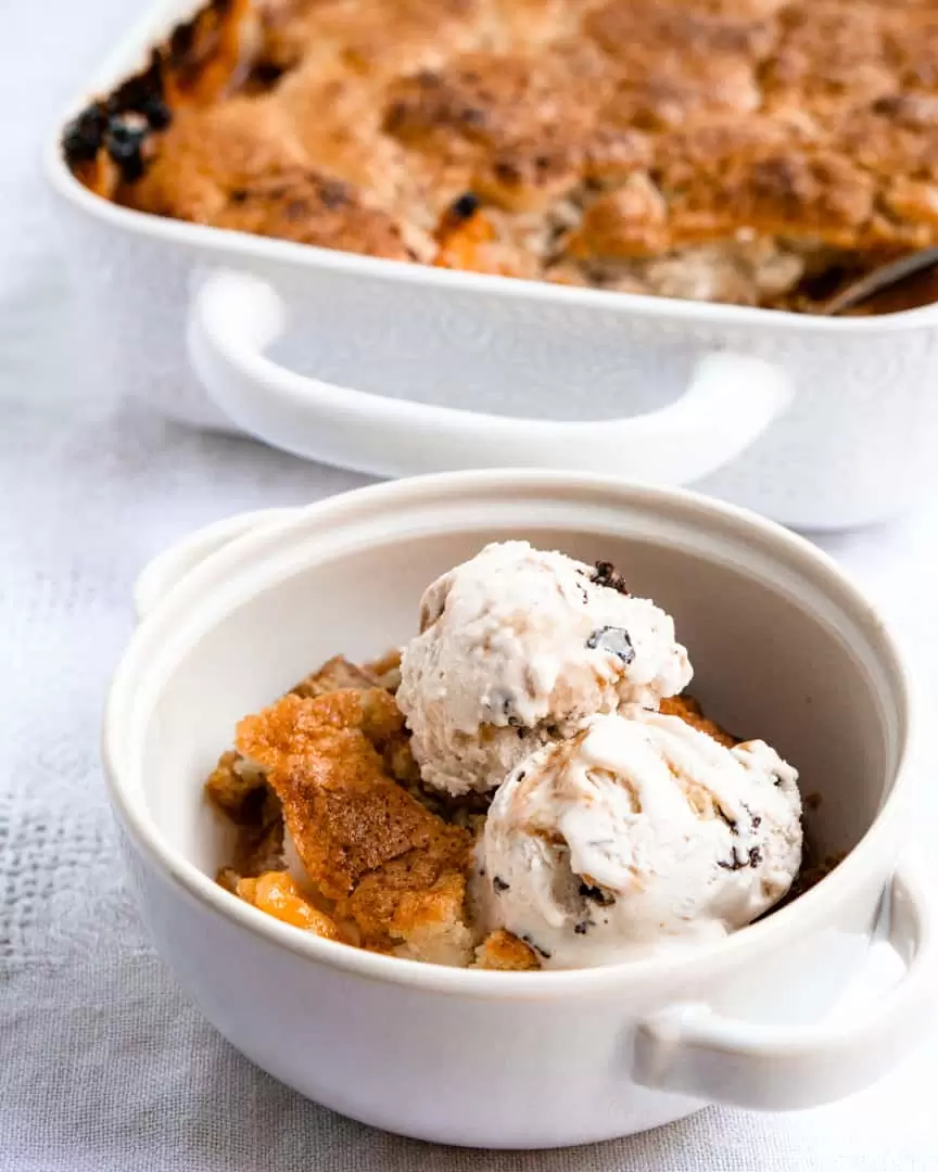 peach cobbler in the background with a small bowl with a scoop of cobbler and two scoops of ice cream on top.