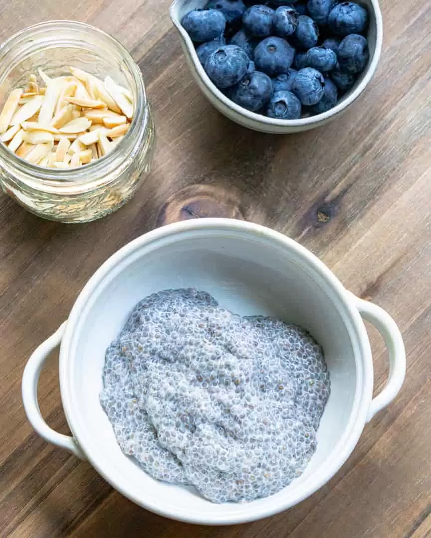 a bowl with chia pudding and two smaller bowls next to it. One has fresh blueberries in it, the other has slivered almonds.