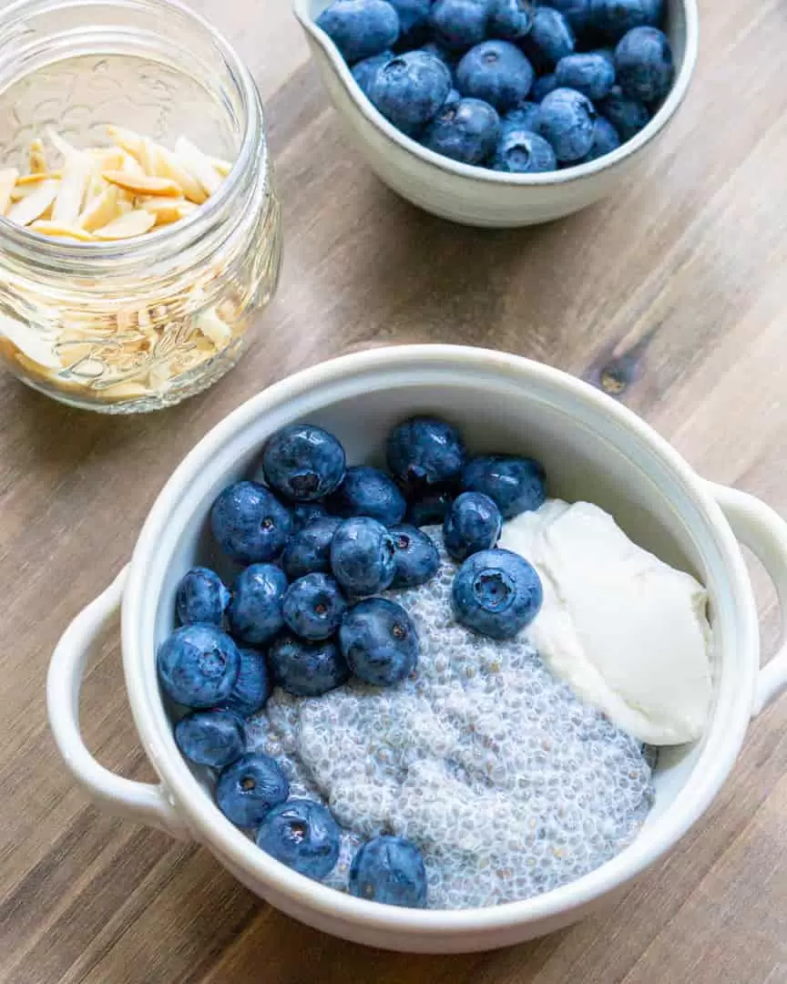 A bowl with chia pudding, yogurt and fresh blueberries. Two other small bowls next to it with fresh blueberries and slivered almonds in them.