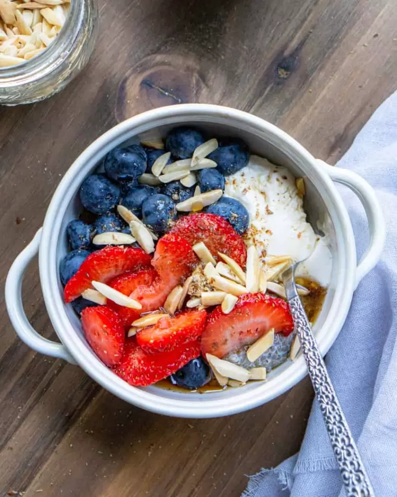 a bowl filled with chia pudding and topped with fresh blueberries, strawberries, yogurt and slivered almonds. The bowl has a spoon in it and a napkin next to it.