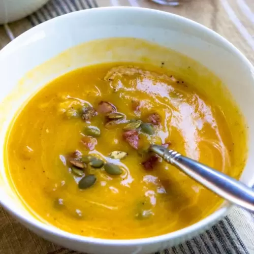 A bowl of butternut squash soup topped with pumpkin seeds and pancetta. There is a spoon in the bowl.