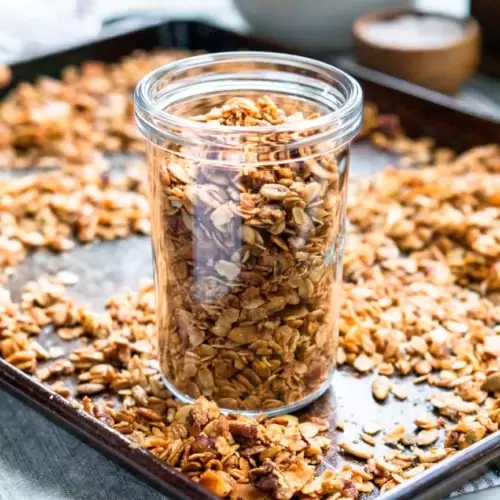 A glass jar filled with granola on top of a baking sheet, surrounded by granola.