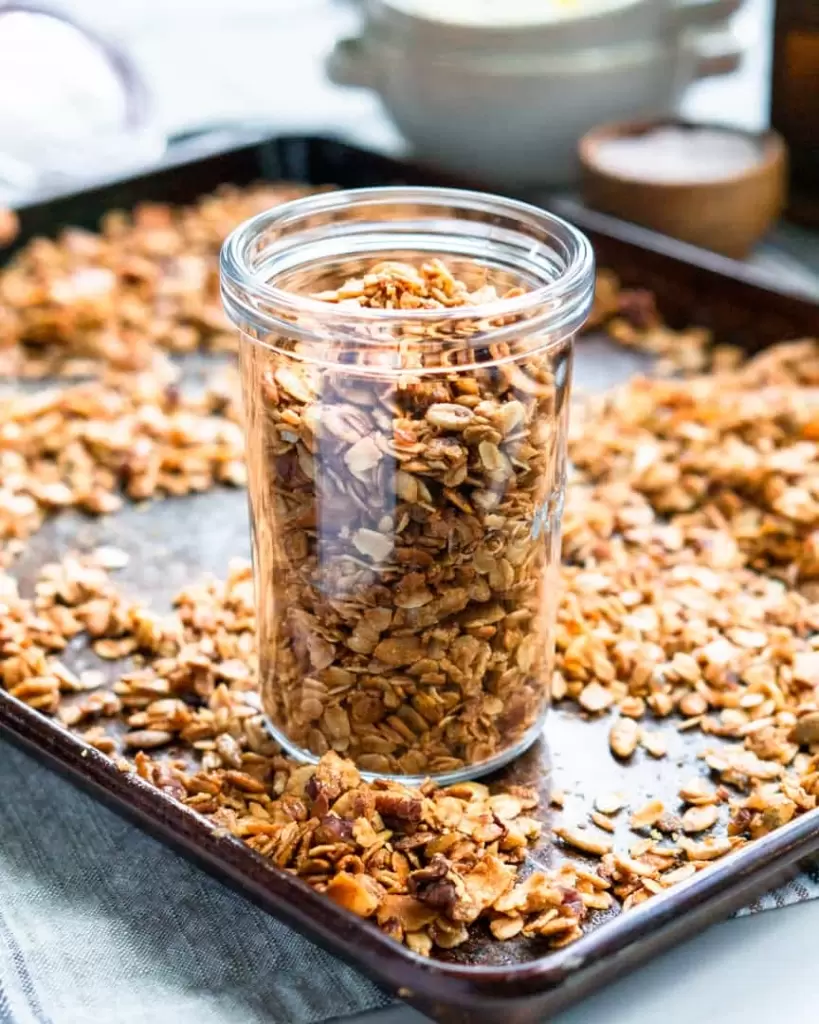 A glass jar filled with granola on top of a baking sheet, surrounded by granola.