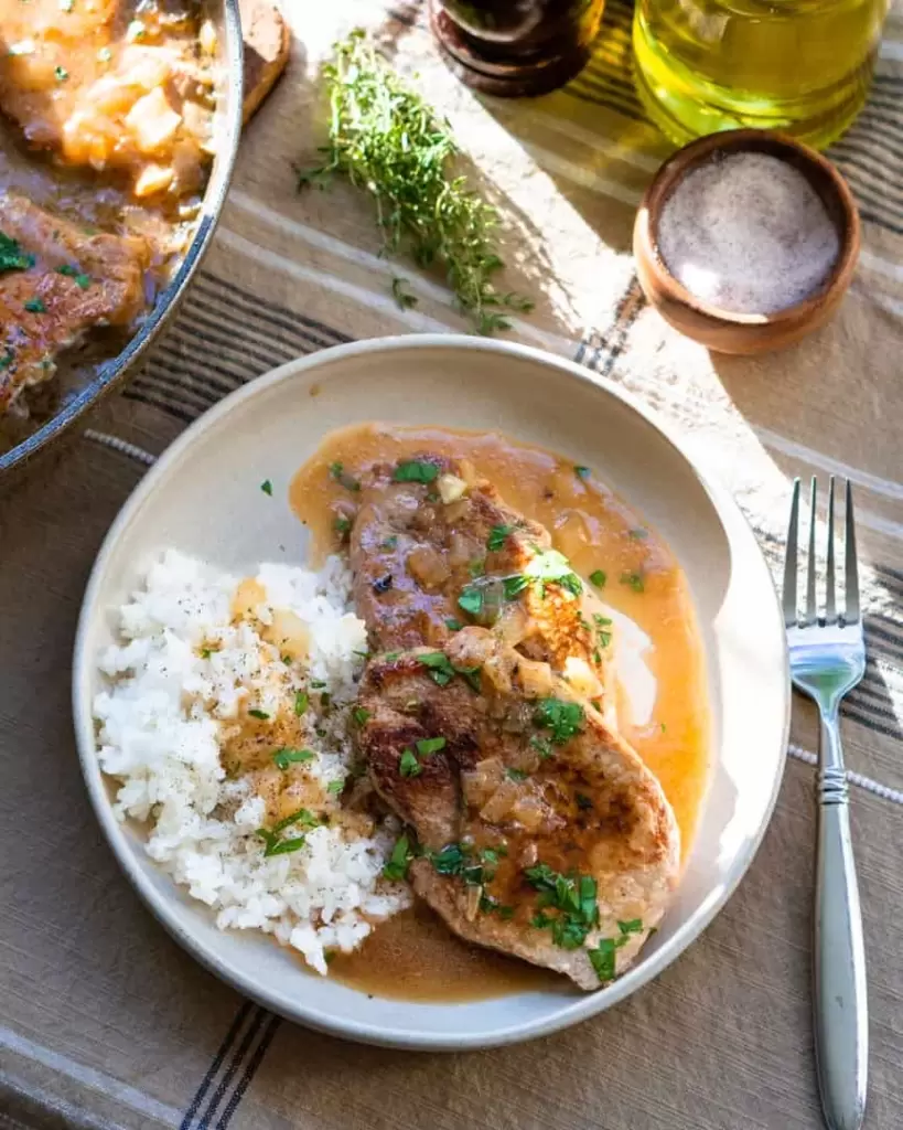 A plate with 2 pork chops with rice next to it. There is pan sauce on the plate and a fork next to it.