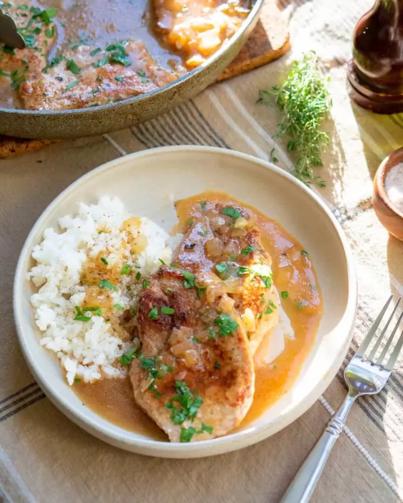 Pork chops on a plate covered in pan sauce with white rice next to it.