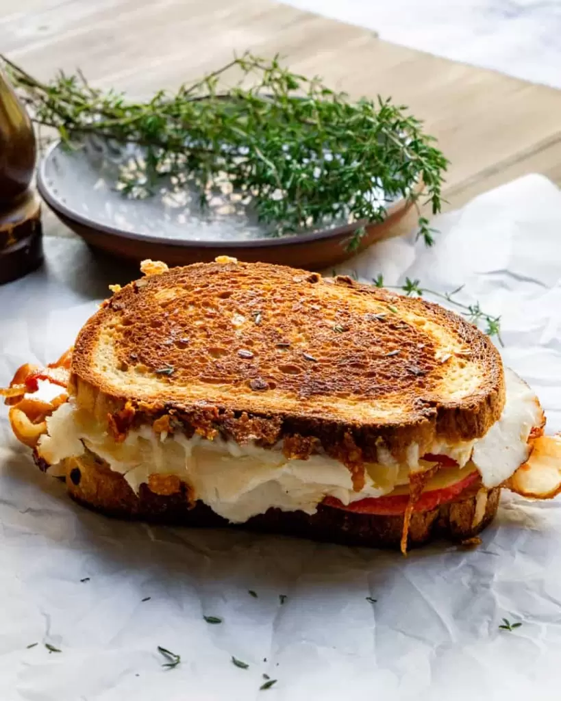 Grilled turkey sandwich with melted cheese and apples on parchment paper. A plate of fresh thyme sitting behind it.