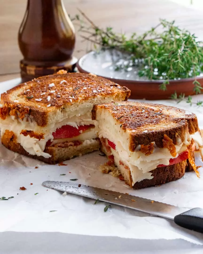 Toasted turkey, apple, bacon sandwich cut in half with a knife placed next to it. A plate of fresh thyme is behind it.