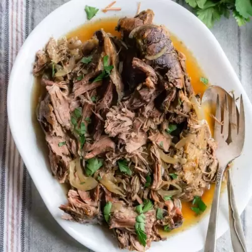 A plate of shredded chuck roast with juices and a fork and spoon for serving also on the plate.