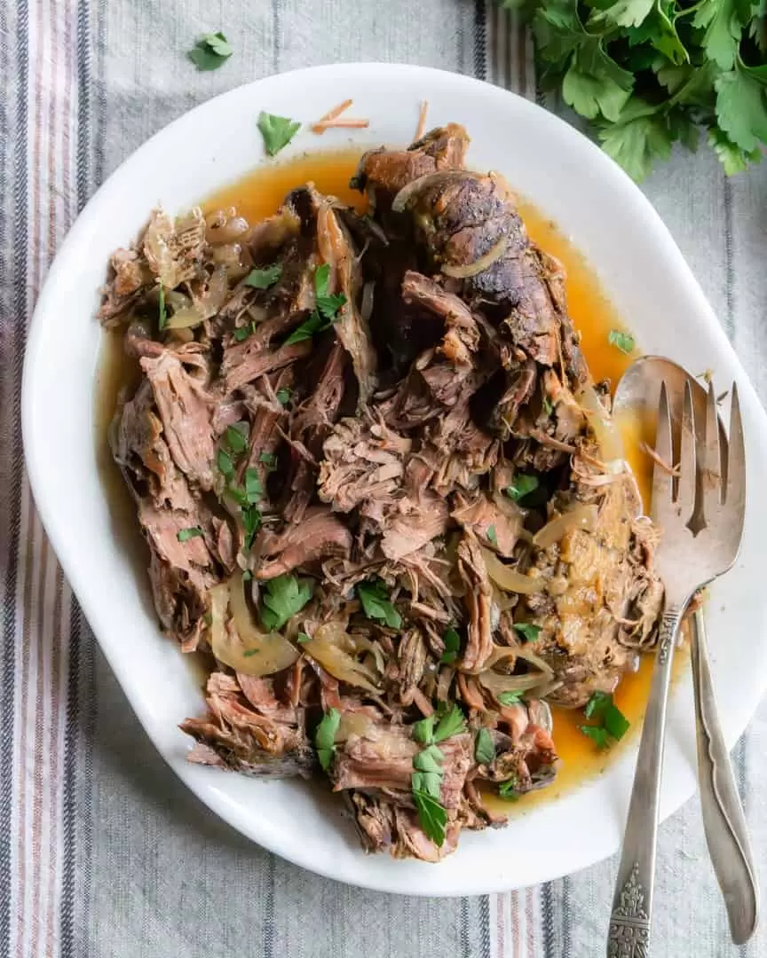 A plate of shredded chuck roast with juices and a fork and spoon for serving also on the plate.