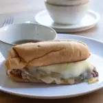 French dip sandwich with melted cheese on a plate with a small bowl of au jus behind it. There are also stacked bowls behind the sandwich.