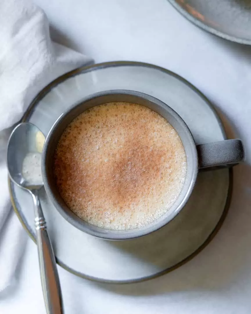 Top down view of a cup of tea on a small plate with a spoon next to it. The tea has a cinnamon sugar topping.