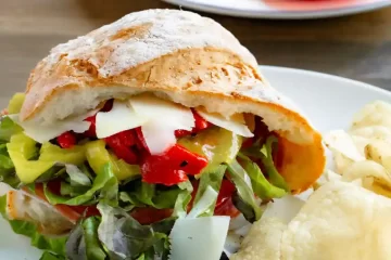 An Italian hoagie on a plate with chips next to it. Sliced tomatoes on a plate in the background