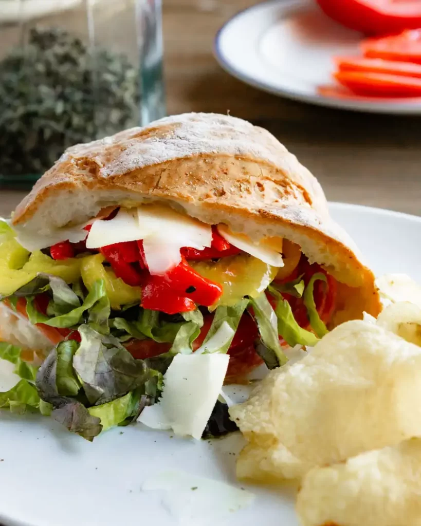 Crusty loaf of bread filled with Italian lunch meats, lettuce, tomatoes and thin shaved slices of provolone cheese. The sandwich is on a plate and there are chips next to it.