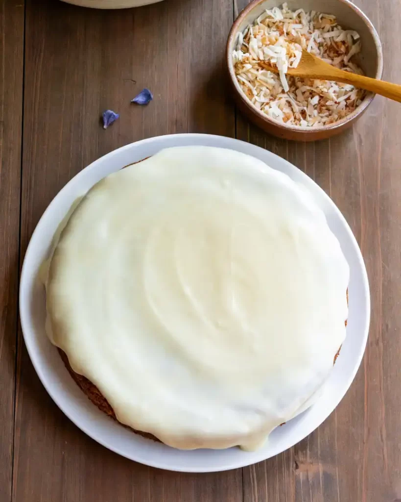 A baked and frosted round carrot cake on a plate. There is a bowl of toasted coconut flakes next to it.