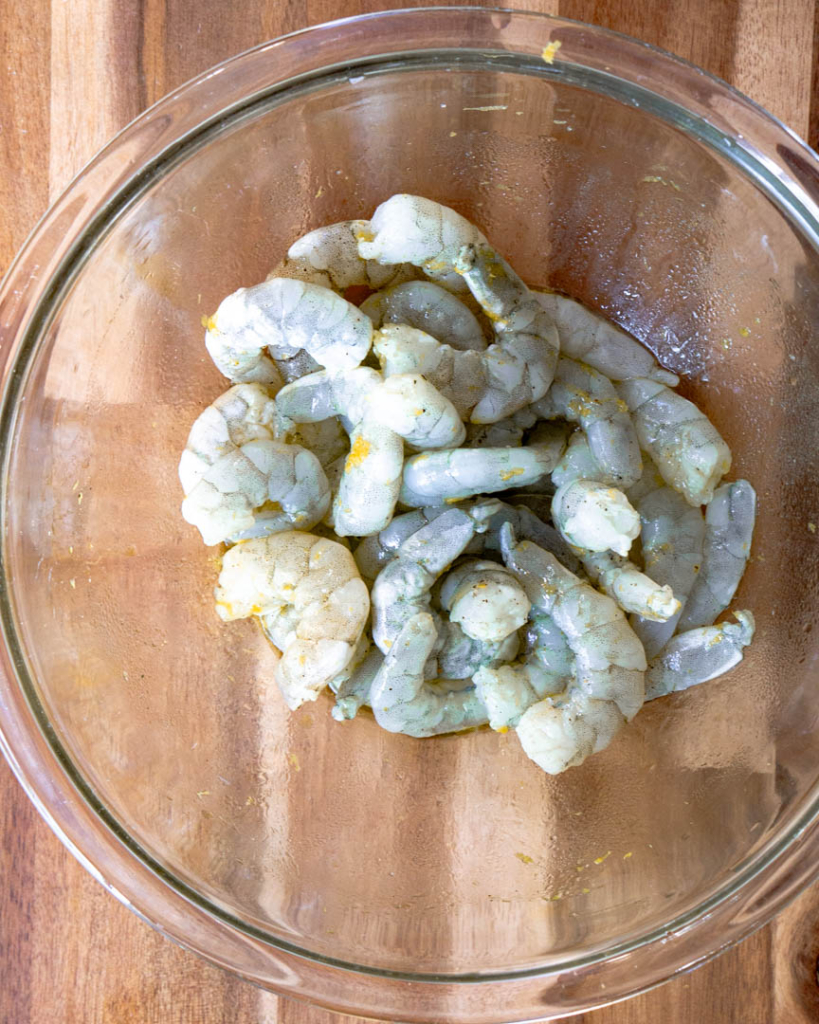 A clear glass bowl with raw shrimp with lemon zest on it.