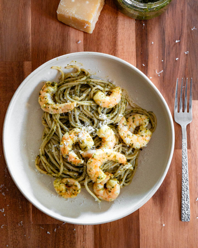 A bowl of spaghetti with pesto and shrimp. There is a fork and a wedge of parmesan cheese next to the bowl.