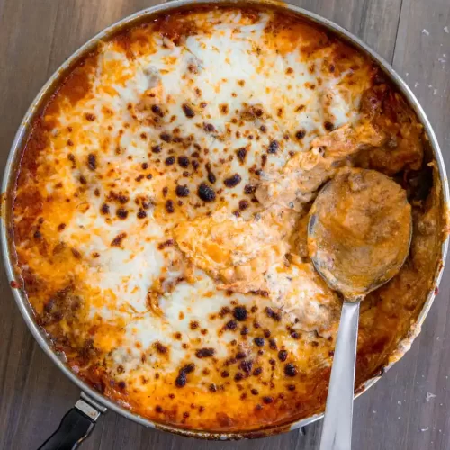 A skillet pan of lasagna with mozzarella that has been broiled. There is a scoop taken out of the pan and a spoon resting in it's place.