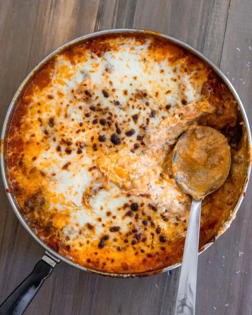 A skillet pan of lasagna with mozzarella that has been broiled. There is a scoop taken out of the pan and a spoon resting in it's place.