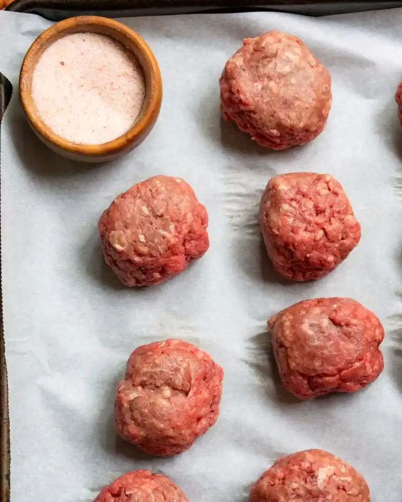 A parchment lined baking sheet with ground beef shaped into balls, ready to be smashed into burgers. There is also a small wooden bowl filled with salt next to the burger balls.