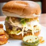 A smash burger with all the toppings and pickles on the plate next to the burger.