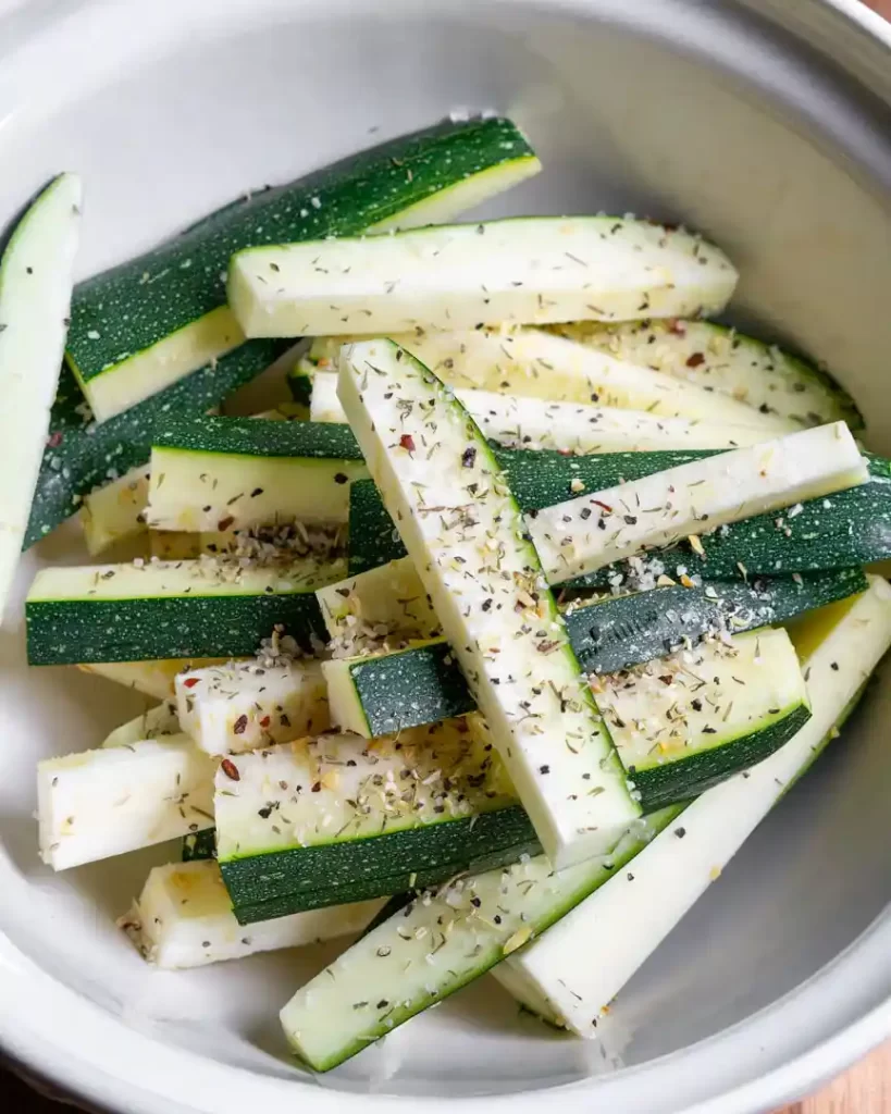 A bowl of raw zucchini spears sprinkled with a steak seasoning.