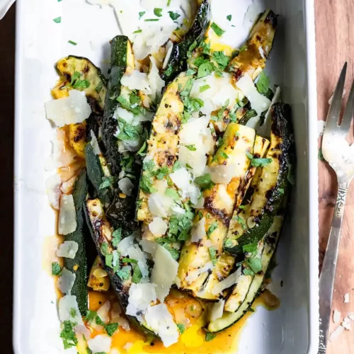 Grilled zucchini spears topped with parmesan cheese shavings and minced fresh parsley in a serving dish.